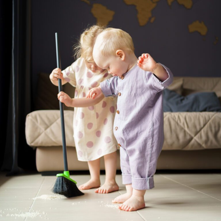 Two small children learning to use a broom to clean up a mess
